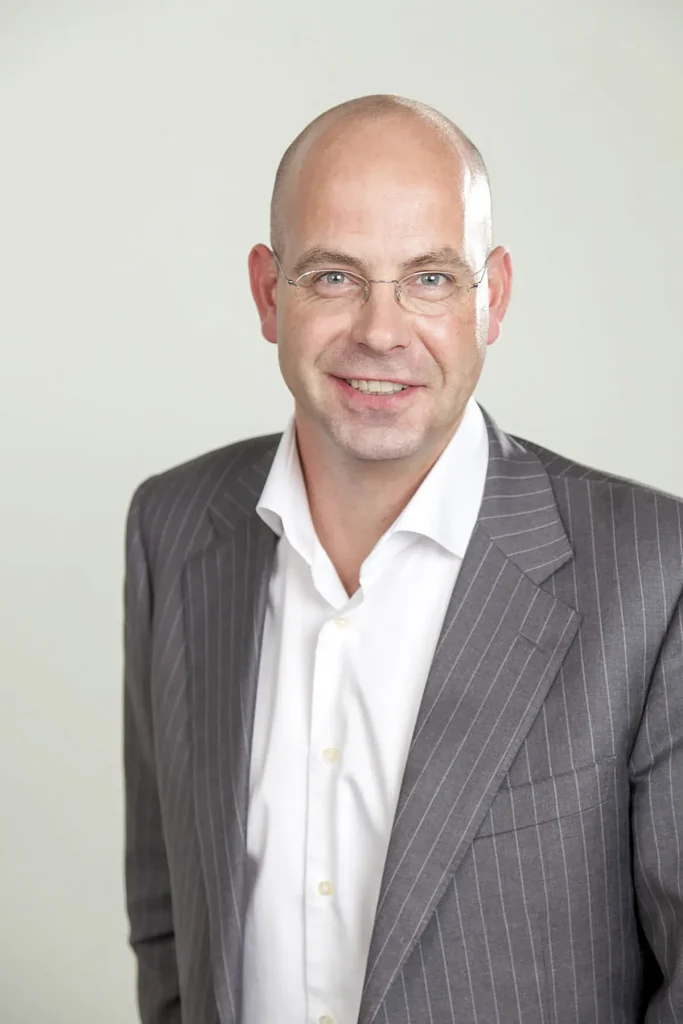 Een kale man met bril lacht naar de camera. Hij draagt een grijs colbert met krijtstreep over een wit overhemd, wat de essentie van bedrijfsfotografie illustreert. De achtergrond is effen gebroken wit en de man straalt een professionele en benaderbare houding uit.