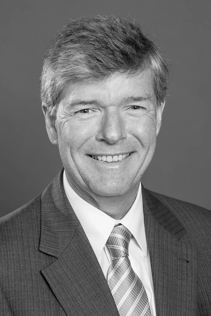 Zwart-wit portret van een man van middelbare leeftijd met kort, netjes gekamd haar. Hij draagt een colbert met krijtstreep, een wit overhemd en een gestreepte stropdas. De man lacht en kijkt recht in de camera, tegen een effen achtergrond in de stijl van bedrijfsfotografie.