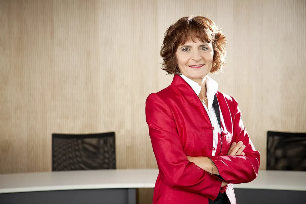 Een vrouw met kort bruin haar, gekleed in een rode blazer over een witte blouse, staat zelfverzekerd met haar armen over elkaar in een moderne kantooromgeving. De achtergrond bestaat uit een houten paneelwand en een wit bureau met zwarte bureaustoelen. Ze lacht en kijkt recht in de camera, perfect voor bedrijfsfotografie.