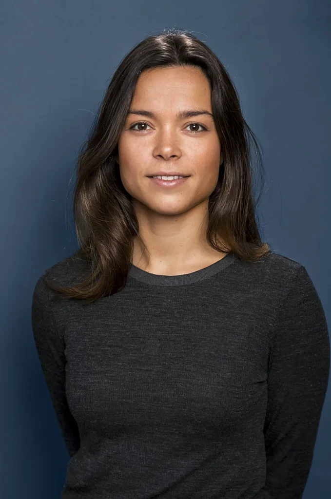 Een jonge vrouw met lang bruin haar en een neutrale uitdrukking staat tegen een effen donkerblauwe achtergrond, een voorbeeld van professionele bedrijfsfotografie. Ze draagt een donkergrijs shirt met lange mouwen en kijkt rechtstreeks in de camera.