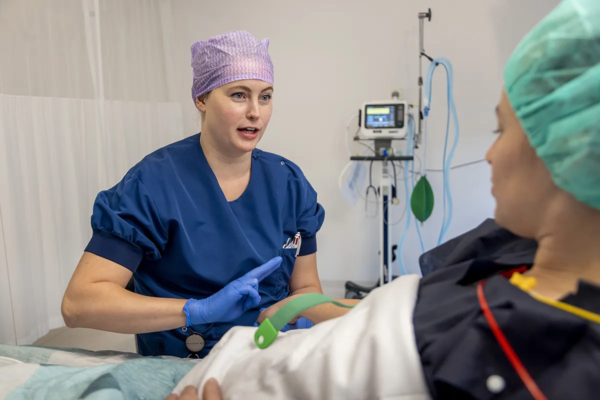 Een verpleegster in een blauw pak en een paarse haarbedekking praat met een patiënt die op een ziekenhuisbed ligt. De patiënt, die een groene operatiemuts en een witte jurk draagt, luistert aandachtig. Op de achtergrond is medische apparatuur zichtbaar, waaronder een monitor met kabels eraan, perfect weergegeven in dit voorbeeld van bedrijfsfotografie.
