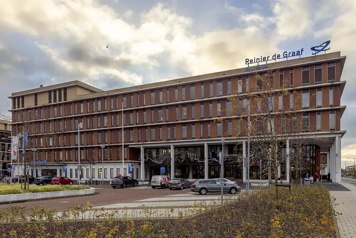 Een groot ziekenhuisgebouw met het opschrift "Reinier de Graaf Gasthuis" heeft een moderne gevel met hoge ramen en metselwerk. Er staan talloze auto's voor de deur geparkeerd en de ingang is versierd met kerstversieringen. De lucht is bewolkt en er zijn kale bomen en struiken op de voorgrond: een perfect tafereel voor bedrijfsfotografie.