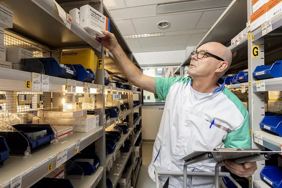 In een goed georganiseerde apotheekopslag haalt een kale man met een bril, een wit en groen uniform en een naamplaatje een medicijndoosje uit een gelabelde plank. Met een klembord en een pen in de hand staat hij tussen netjes geordende blauwe bakken en diverse medicijnen, warm verlicht door de verlichting. Ideaal voor bedrijfsfotografie.
