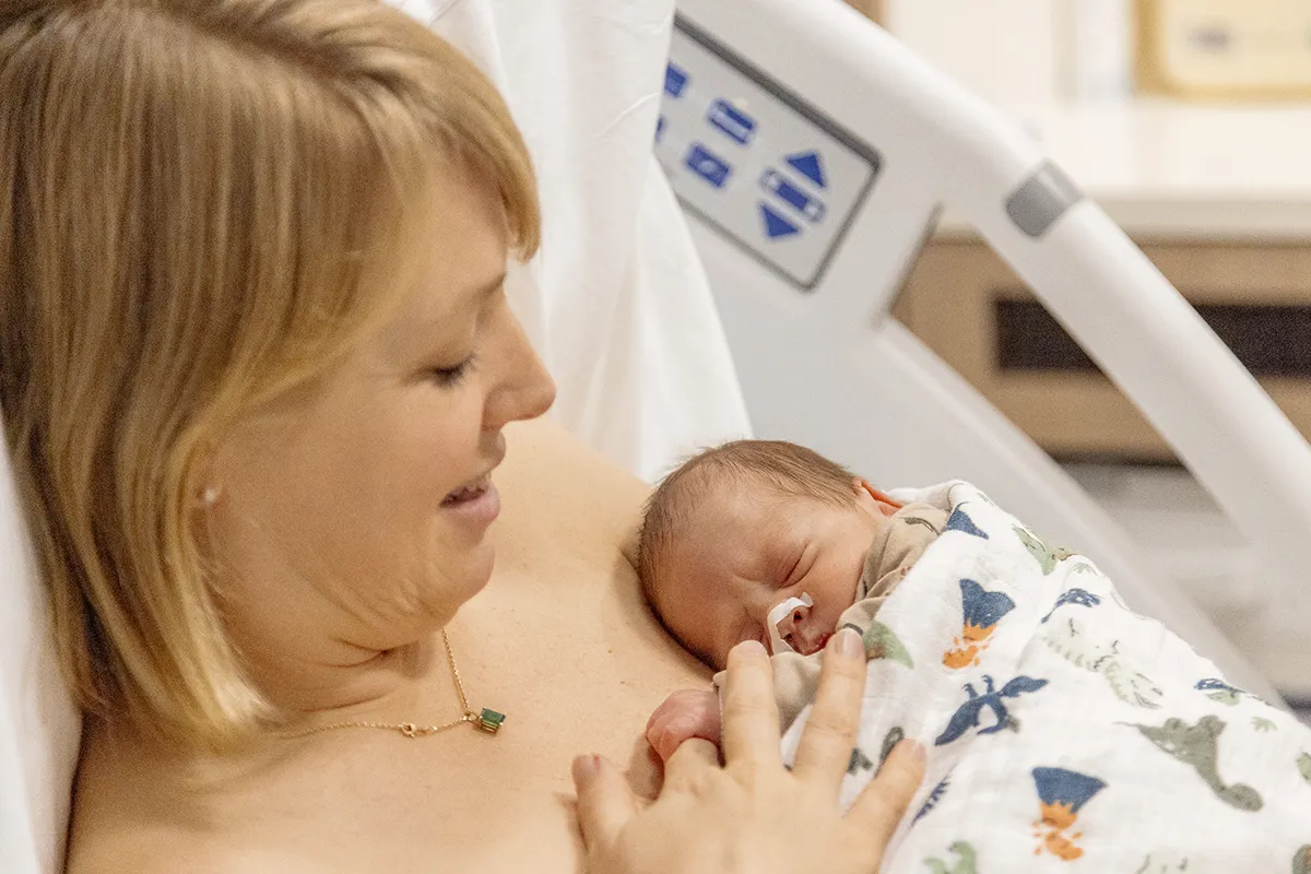 Een vrouw met kort blond haar glimlacht terwijl ze een pasgeboren baby vasthoudt, gewikkeld in een deken met patronen. Ze ligt in een ziekenhuisbed en draagt een ziekenhuisjas. De baby heeft een voedingssonde in de neus en ligt vredig tegen de borst van de vrouw te slapen, terwijl ziekenhuisapparatuur op de achtergrond zichtbaar is, waardoor dit intieme moment wordt vastgelegd dat doet denken aan oprechte bedrijfsfotografie.