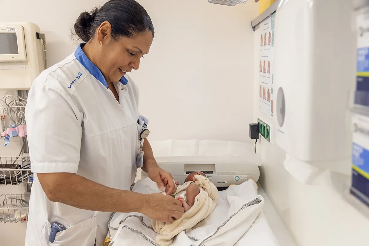 Een verpleegster in een wit uniform verzorgt een pasgeboren baby, gewikkeld in een crèmekleurige deken op een commode in een ziekenhuiskamer. Medische apparatuur en kaarten zijn zichtbaar aan de muur, wat een sfeer benadrukt die perfect is voor bedrijfsfotografie. De verpleegster glimlacht en concentreert zich op de baby, waardoor een verzorgende en aandachtige scène ontstaat.