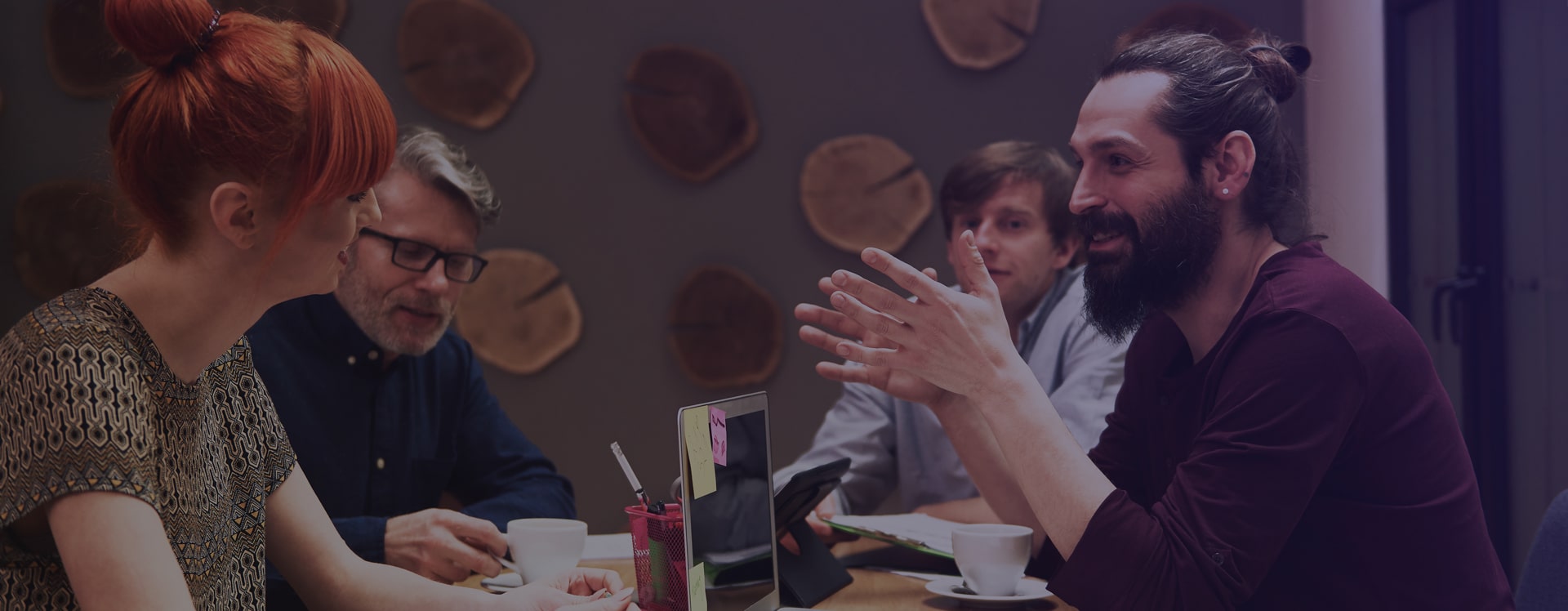 Vier collega's in levendig overleg aan een vergadertafel met laptop en koffie, warm donker interieur met houten wanddecoratie